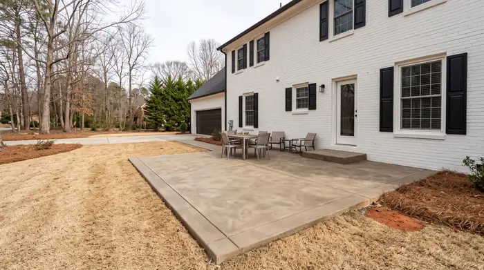 Rectangular concrete patio with a low step up to the rear door at a painted-white-brick two-story home in Lawrenceville, GA, under bright midday cloud.
