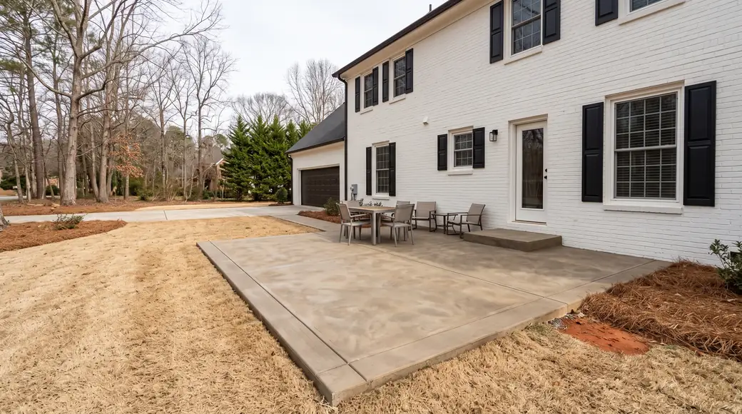 Rectangular concrete patio with a low step up to the rear door at a painted-white-brick two-story home in Lawrenceville, GA, under bright midday cloud.