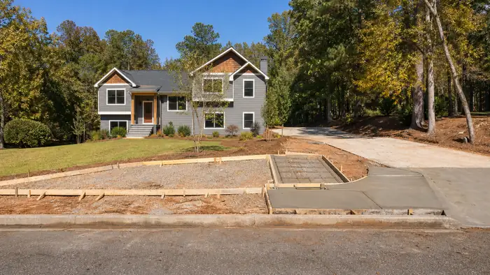 New concrete sidewalk being formed along a street with a driveway ramp at a grey split-level home in Lawrenceville, GA, in clear late-morning sun.